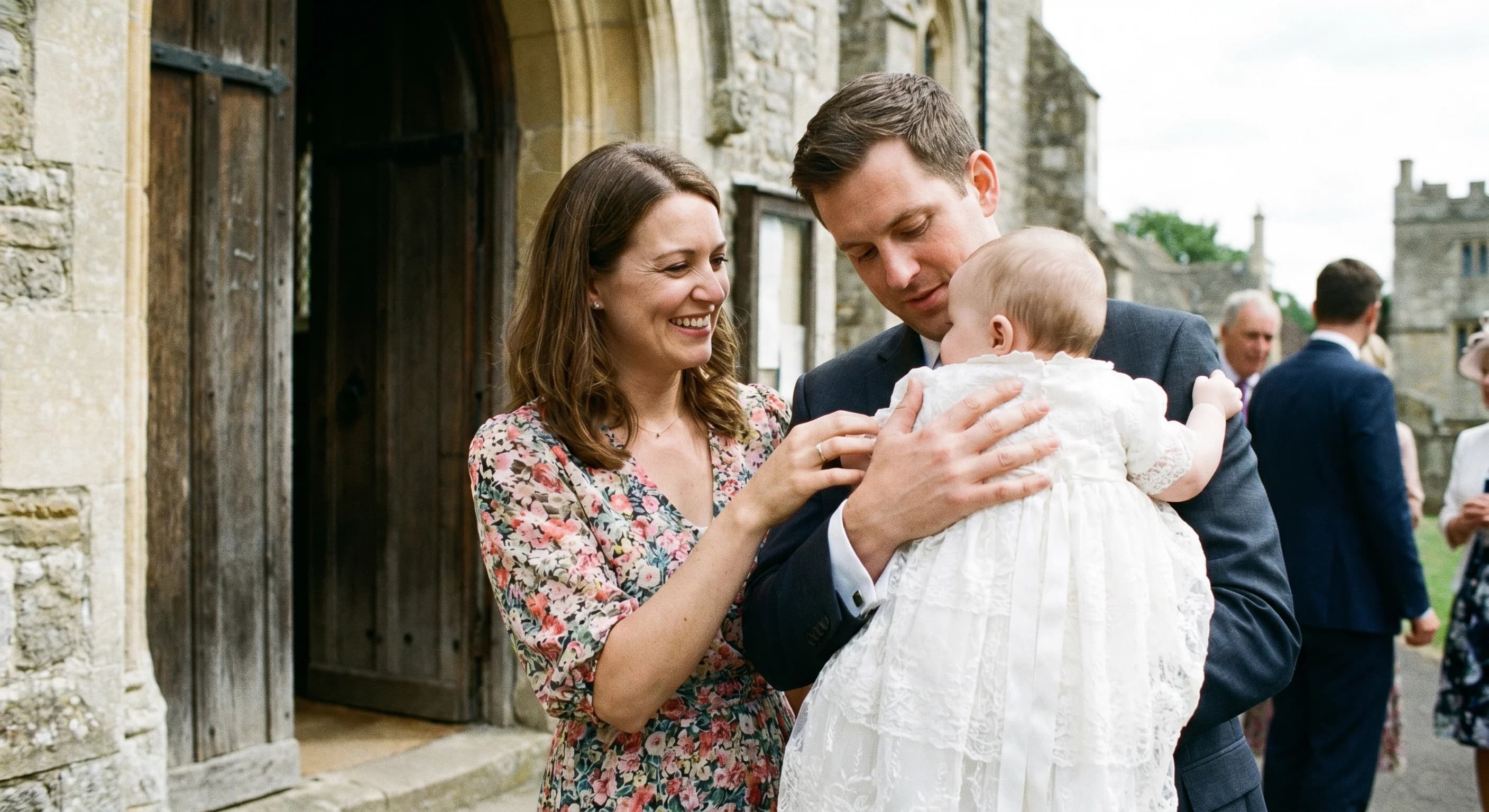 Two godparents holding a baby in a white christening gown outside a church entrance