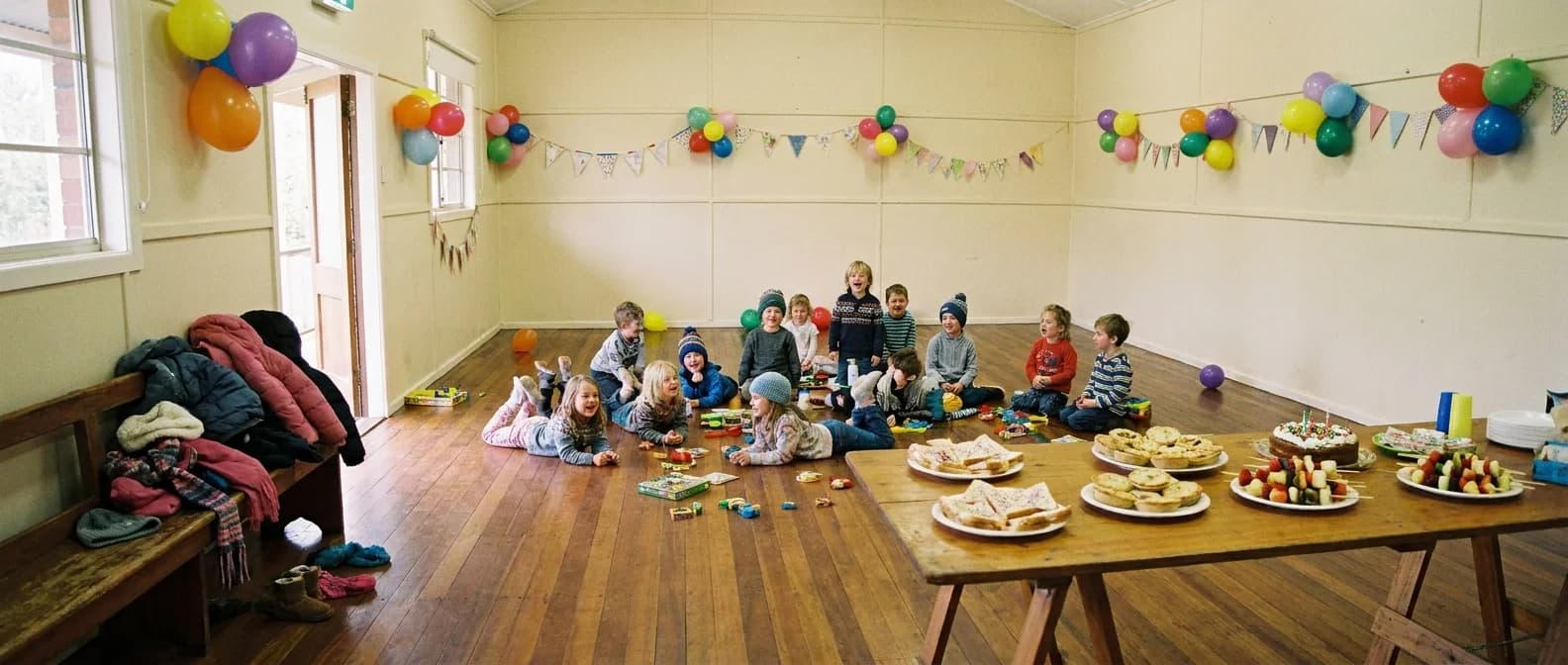 Children mid-laugh at a school-holiday birthday party inside a bright community hall, balloon clusters and bunting on the walls, a table of party food in the foreground, coats and beanies piled on a bench
