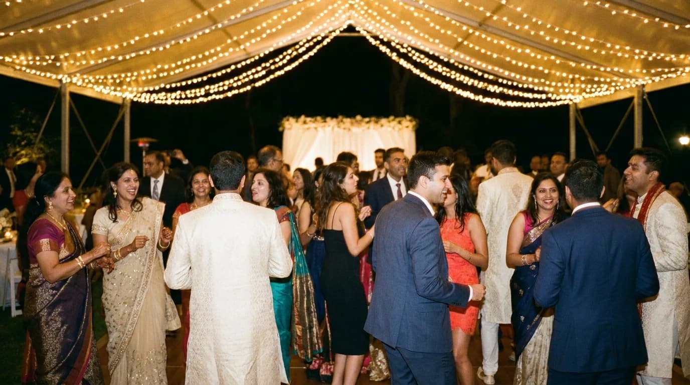 Guests in a mix of Indian formalwear and cocktail attire dancing together under string lights at a wedding reception